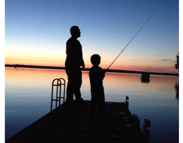 A boy fishing with dad on a dock at sunset.